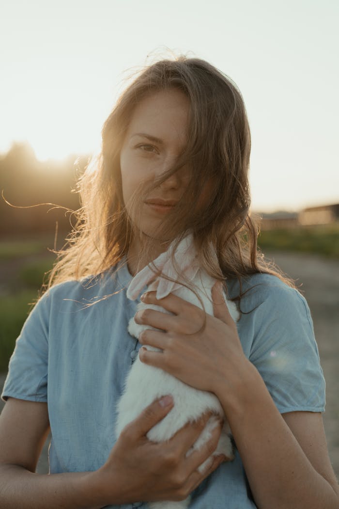 services-03 A young woman embraces a white rabbit during a serene sunset in an open field.